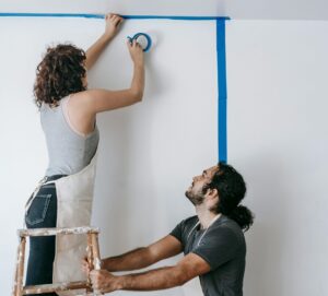 Photo of a man and woman preparing to paint a wall. The man holds a ladder while the woman stands on the ladder and puts painter's tape up