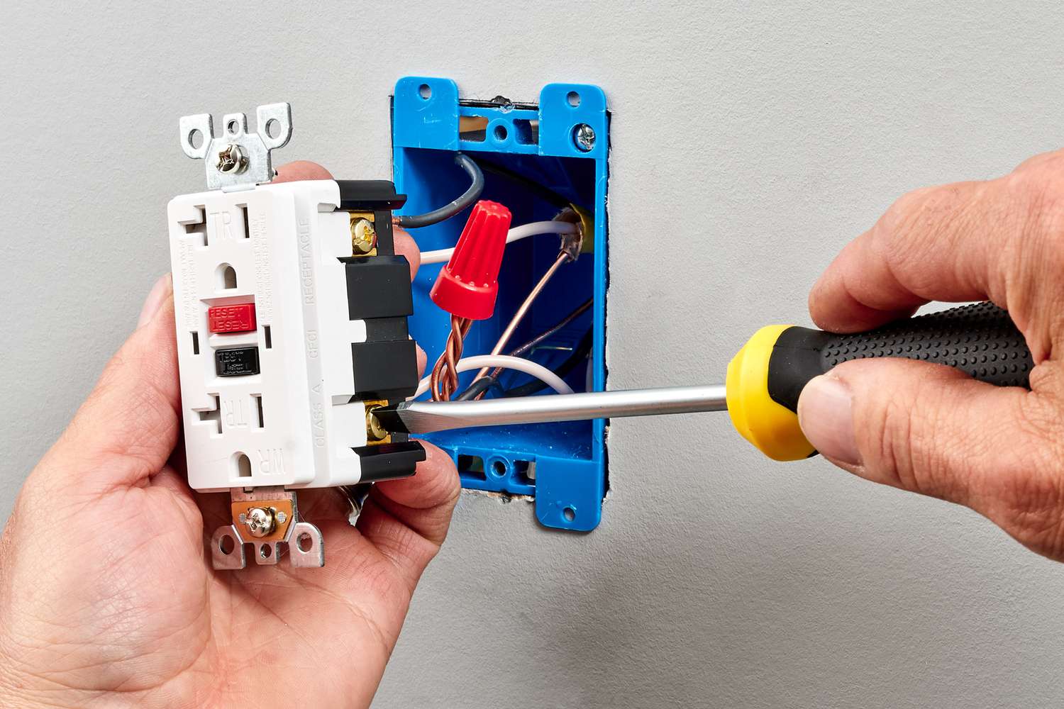 Close up photo of two hands, one holding a wall outlet that has been removed to expose the electrical wiring, one holding a screwdriver